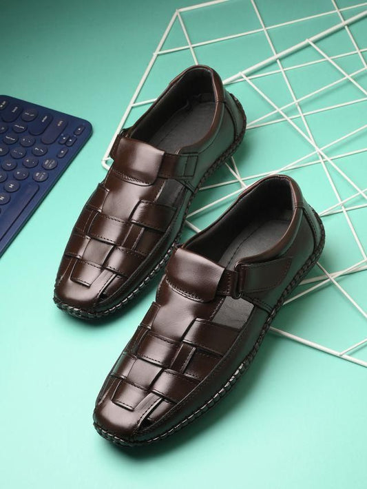 Elegant brown leather men's slippers with a woven pattern, resting on a mint green and white grid surface alongside a calculator.